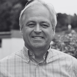 A grayscale photo of a person with short, light-colored hair, wearing a plaid button-up shirt. They are smiling and standing outside, with greenery and a blurred building in the background.