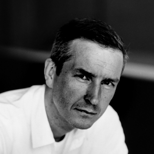 Black and white portrait of a man with short hair, wearing a white shirt. He gazes directly at the camera, with a neutral expression. The background is blurred, focusing attention on the subject's face.