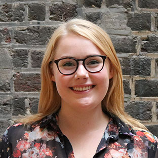 A person with long blond hair and glasses smiles at the camera. They are wearing a floral-patterned blouse. The background features a textured stone wall.