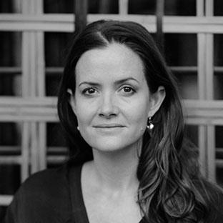 A black and white portrait of a woman with long hair, wearing earrings and a dark top. She is looking directly at the camera, standing in front of a lattice-style background.