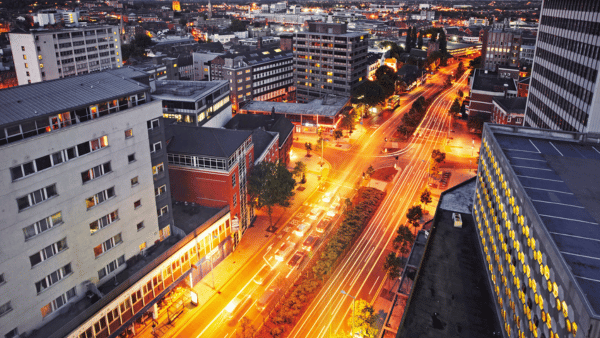 Aerial view of a city at night with illuminated streets and light trails from moving vehicles. Tall buildings line the brightly lit roads, and the cityscape extends into the distance under a dark sky.
