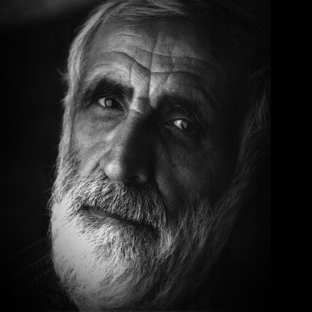 Black and white portrait of an older man with a thick white beard and expressive eyes, looking slightly upward. The lighting highlights the texture of his skin and hair, creating a reflective and thoughtful mood.
