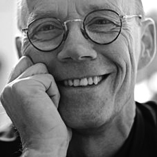 A black and white close-up of a smiling person wearing round glasses, resting their face on their hand. The image captures the subject's friendly expression and relaxed demeanor.