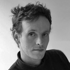 A black and white portrait of a person with short, slightly tousled hair, looking at the camera. The person is wearing a dark, high-collared shirt. The background is a plain, light color.