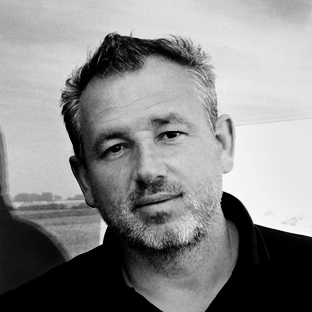 Black and white portrait of a man with short hair and a beard, wearing a collared shirt. He looks directly at the camera with a neutral expression. The background is a blurred landscape.