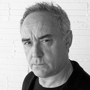 Black and white portrait of a person with short, curly hair, wearing a dark jacket over a black shirt. They are in front of a white brick wall, looking directly at the camera with a serious expression.