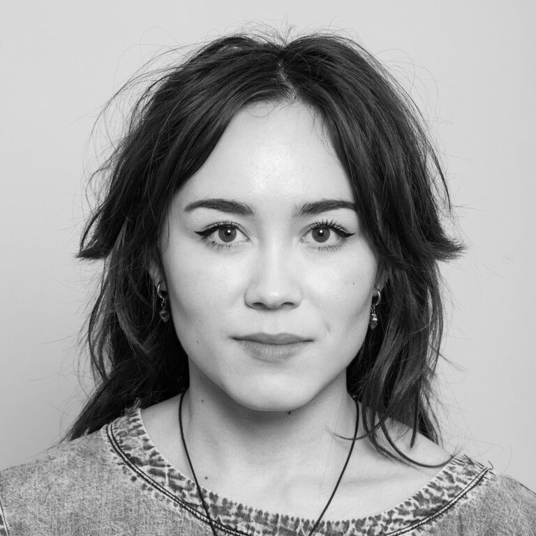 Black and white portrait of Fionna Monk with long dark hair and a relaxed expression, wearing a textured top and a necklace, looking directly at the camera against a plain background.