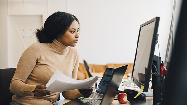 A person wearing a tan turtleneck is sitting at a desk, focused on a desktop computer screen. They hold papers in one hand and have an open laptop beside them. A bitten apple rests on the desk, and the background features office seating.