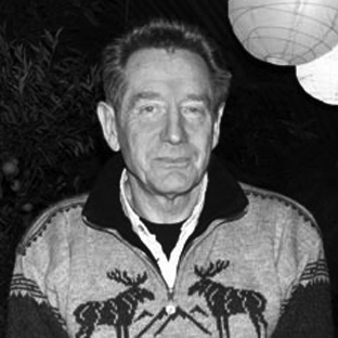A grayscale photo of a man wearing a patterned sweater with reindeer designs. He stands outdoors at night, with foliage and a hanging paper lantern in the background.