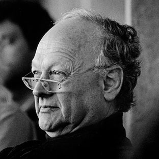 Black and white image of an older man with glasses resting on his nose. He has a serious expression and is looking to the side. The background is blurred, with another person visible out of focus.