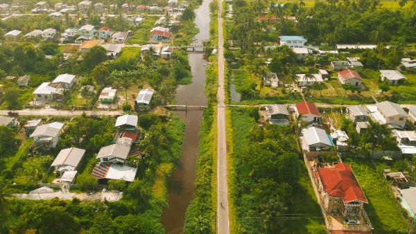 Aerial view of a village with houses on either side of a narrow canal. The landscape is lush and green, and a straight road runs parallel to the canal, crossing it via a small bridge. The rooftops display a mix of colors and materials.