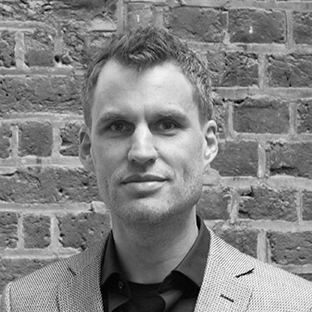 Black and white photo of a man with short hair, wearing a light-colored blazer over a dark shirt, standing in front of a brick wall.