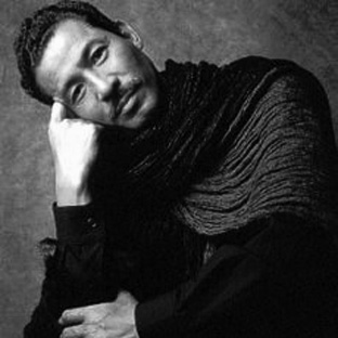 Black and white portrait of a person with short curly hair, wearing a textured, dark scarf over a dark shirt. The person is resting their head on their hand, looking thoughtfully at the camera against a textured background.