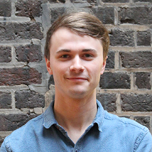 A person with short light brown hair smiles subtly, wearing a denim shirt. They are standing in front of a textured brick wall.