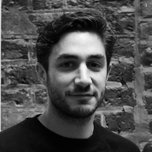 A person with short, dark hair and a beard is standing in front of a brick wall, wearing a dark shirt. The photo is in black and white.