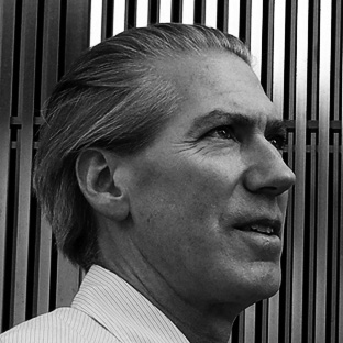 Black and white photo of a man with light-colored hair, wearing a collared shirt, looking to the right. Vertical bars form the background.