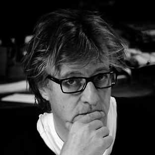 Black and white photo of a person with medium-length hair and glasses, resting their chin on their hand. The background is out of focus, highlighting the contemplative expression on their face.