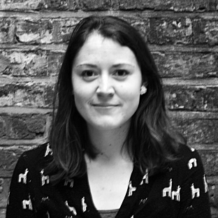 Black and white portrait of a person with shoulder-length hair, smiling slightly. They are wearing a dark sweater with patterns in front of a brick wall background.