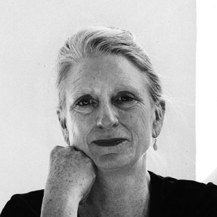 Black and white photo of a person with their chin resting on their hand. They have light hair pulled back and are wearing a dark top. The background is plain.