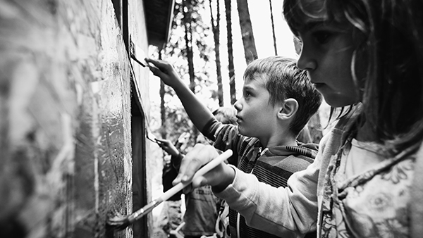 Black-and-white image of two children focused on painting a wall with brushes. The boy in the middle wears a striped hoodie; the girl on the right, a patterned jacket. The background shows blurred trees and vertical beams.