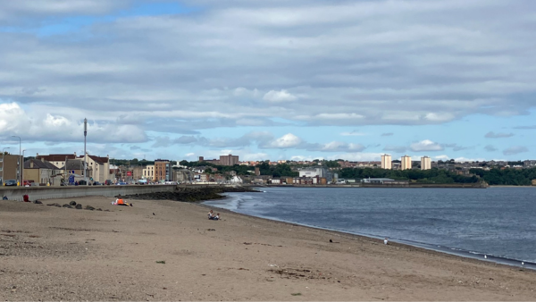 A sandy beach with a few people sitting scattered along its length. The shoreline curves gently, leading to a nearby town. Buildings and greenery are in the background, under a cloudy sky.