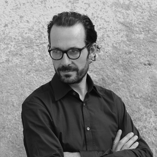 A man with glasses and a beard stands against a textured wall, wearing a dark button-up shirt. His arms are crossed, and he looks thoughtfully to the side. The image is in black and white.