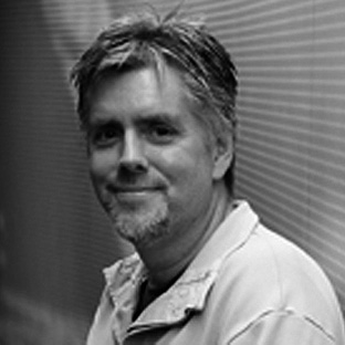 A grayscale image of a man with short hair and a beard, smiling slightly. He is wearing a collared shirt and standing against a textured background. The lighting creates subtle contrasts on his face and clothing.