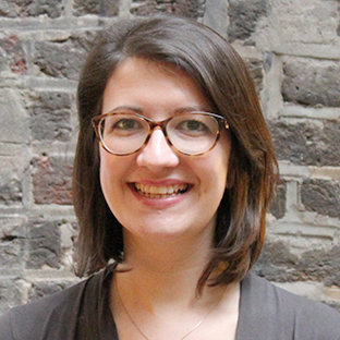 A person with shoulder-length brown hair and glasses smiles at the camera. They are wearing a dark top and standing in front of a brick wall.