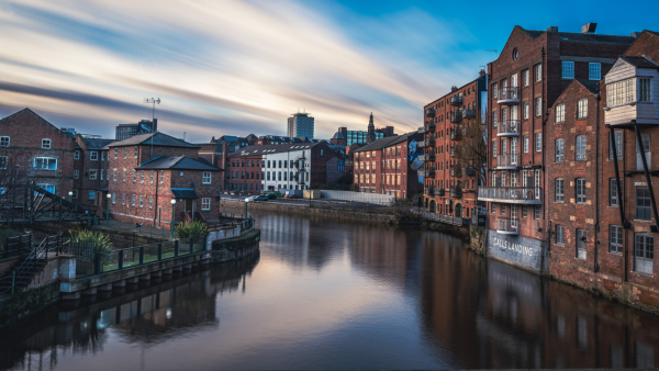 A tranquil canal scene in a city with red-brick buildings on both sides, reflected in the calm water. The sky is a blend of blue and white due to long exposure, creating a smooth, dreamy effect. Urban architecture lines the riverside.