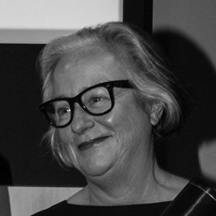 A person with short, light hair and black-framed glasses smiles while looking slightly to the side. The image is in black and white, showing them from the shoulders up.