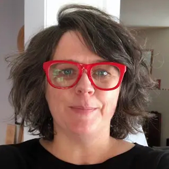 Person with shoulder-length dark hair wearing large red glasses and a black shirt, standing indoors.