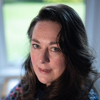 A person with long, dark hair and a patterned top is looking at the camera. The background features a softly focused view of a green lawn seen through tall windows.