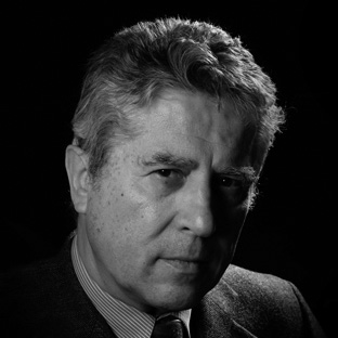 Black and white portrait of an older man with wavy hair, wearing a suit. He looks directly at the camera against a dark background, with an expression of seriousness.