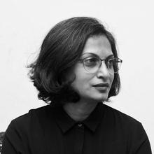 Black and white portrait of a person with medium-length dark hair and glasses, wearing a dark collared shirt, looking to the side against a plain light background.