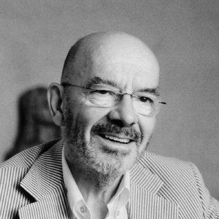 A black and white portrait of a smiling man with glasses and a short beard. He is wearing a striped jacket over a white shirt. The background is plain and blurred, focusing attention on the man's face.
