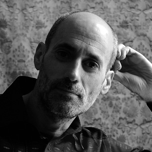 Black and white portrait of a man with a beard, wearing a patterned dark shirt. He is resting his head on his hand, looking toward the camera, against a textured background.
