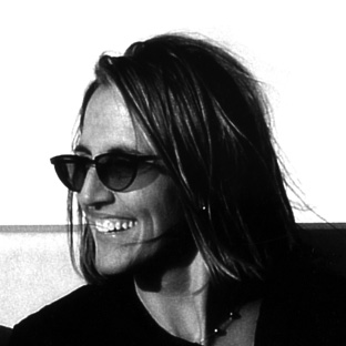 A person with long hair wearing sunglasses is smiling while looking to the side. The black and white photo captures them against a bright background.