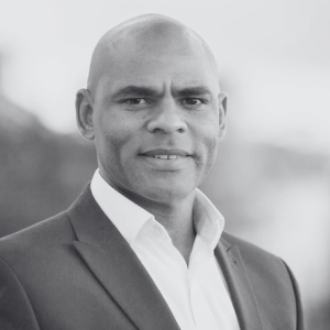 A man in a suit and open-collar shirt smiles slightly while looking at the camera. The background is blurred, suggesting an outdoor setting. The image is in black and white, highlighting his facial features and attire.