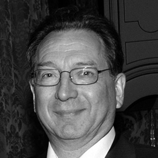 A black and white photo of a man wearing glasses and a suit. He is smiling slightly. The background features patterned wallpaper and ornate wood detailing.