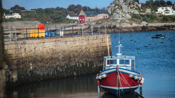 A red and white boat docked by a stone wall in a small harbor. Behind, there is a rocky landscape with houses and green trees. The water is calm with some buoys visible.