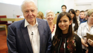 An older man in a blue suit stands beside a young woman with long dark hair, wearing a black floral jacket. They are in a room with a crowd of people smiling in the background.