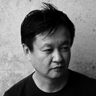 A black and white portrait of a person with short, dark hair, looking down and to the side. They wear a dark shirt and are positioned against a textured wall.