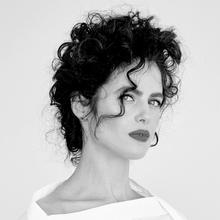 Black and white portrait of a person with curly hair styled up, wearing a white top. They have a serious expression and are looking slightly to the side.