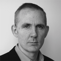 A black and white portrait of a person with short hair, wearing a suit jacket over a collared shirt. They are facing the camera with a neutral expression, and the background is a plain white wall.
