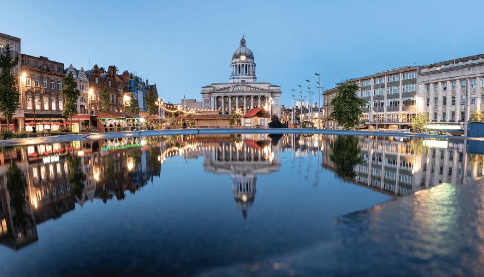 A large, dome-topped building with columns is reflected in a calm pool of water during twilight. The scene is surrounded by lit buildings and streetlights, creating a serene urban atmosphere.