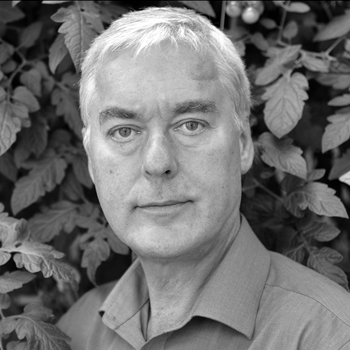 A grayscale portrait of a person with short white hair, wearing a collared shirt, standing in front of leafy plants. Their expression is neutral, looking directly at the camera.