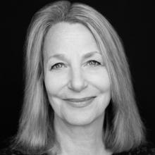 A black and white photograph of a woman with medium-length hair. She is smiling softly and looking directly at the camera. The background is a solid black, highlighting her expression.