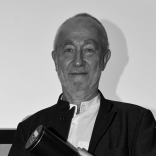Elderly person with a short beard holding a cylindrical object, wearing a dark jacket over a light shirt, smiles at the camera. Black and white photograph with a plain background.