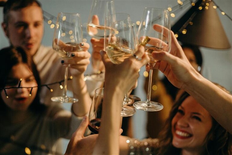A group of people cheerfully clinking glasses at a celebration, surrounded by string lights. Some hold wine glasses filled with drinks, and one person holds a glass of red wine. The atmosphere is festive and joyful.