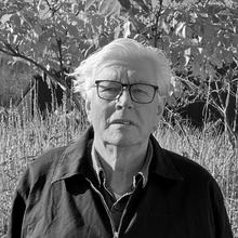 An older person with glasses stands outdoors in front of tall grasses and trees. The image is in black and white.